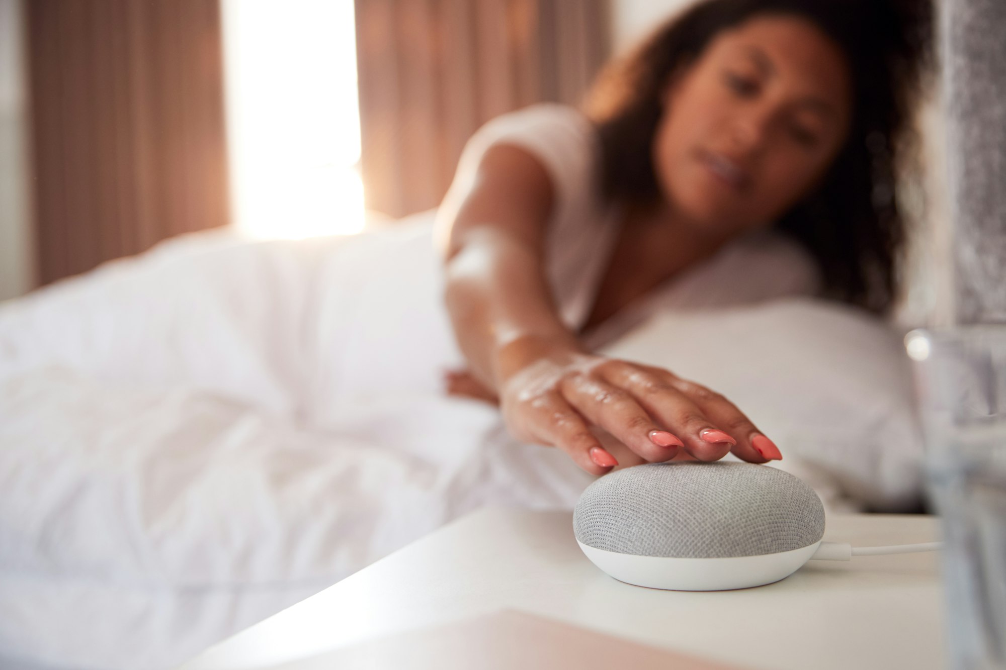 Woman Waking Up In Bed With Voice Assistant On Bedside Table Next To Her